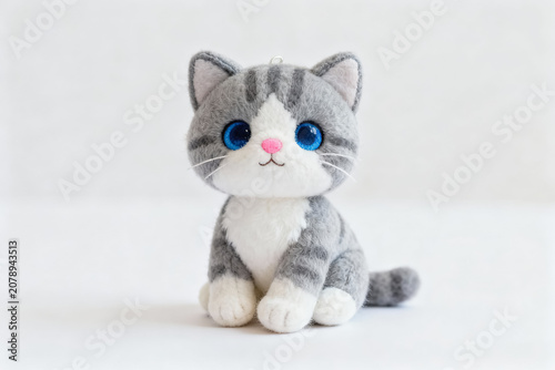 A small, soft plush kitten toy with gray and white fur, big blue eyes, and a pink nose, sitting on a plain white surface.