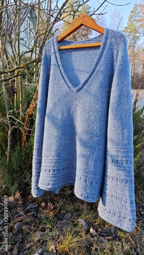 Light blue knitted V-neck sweater hanging on a wooden hanger outdoors. Handmade warm winter clothing