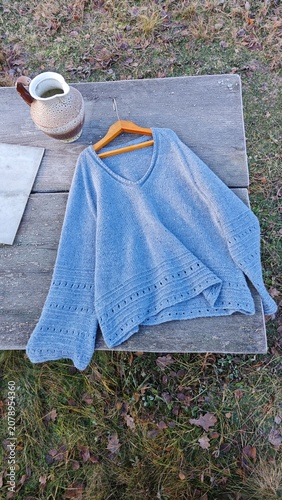 Top view of a light blue knitted sweater and ceramic pitcher on a wooden table. Handmade v-neck knitwear outdoors