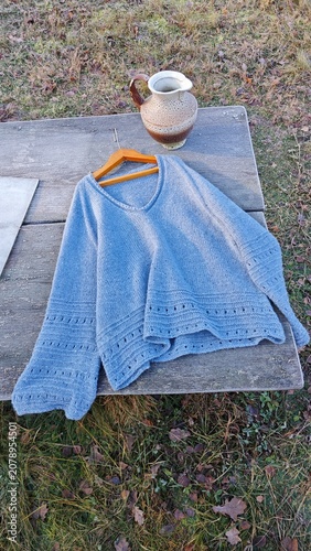 Flat lay of a light blue knitted sweater and ceramic pitcher on a wooden table. Handmade winter clothing outdoors