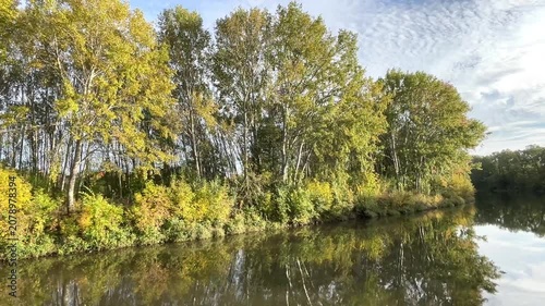 calm river, yellow-green leaves on the trees, an idyllic river landscape seen from a boat in autumn, sunny day, blue sky, church in the background, trees reflected in the river, green reflection