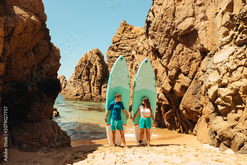 beach Ursa, Portugal. Couple holding hands on beach with surfboards near rock formations in bright sunlight.  They near tall rock formations and each holds a surfboard.  sun shines brightly above them