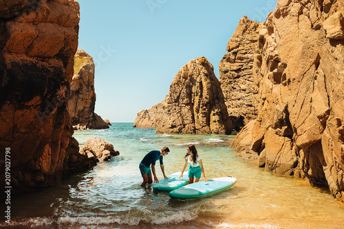 beach Ursa, Portugal. Two people prepare stand-up paddle boards near rocky shore during bright day. sup boards on sandy beach by water surrounded by large rocks under clear blue sky in midday sun.