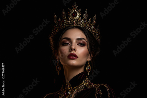 A beautiful woman wearing an elaborate golden crown and regal attire is posing against a black background.