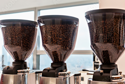 Coffee. Three coffee grinders are positioned in row, each featuring transparent hopper filled with roasted coffee beans. Background shows bright cafe interior with large windows