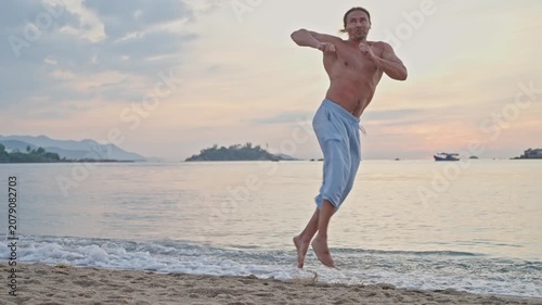 Shirtless martial artist practicing high fly kick on sandy beach at golden sunset, facing calm sea with distant islands, small boat, gentle waves, warm backlight, focused balanced