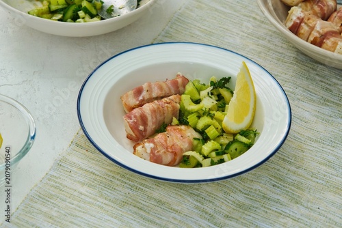 Grilled cod wrapped in bacon strips with celery and cucumber salad on a white plate against a white concrete background.