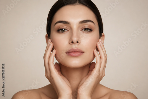 A young woman with brown hair is posing and gently touching her cheeks against a plain, light background.