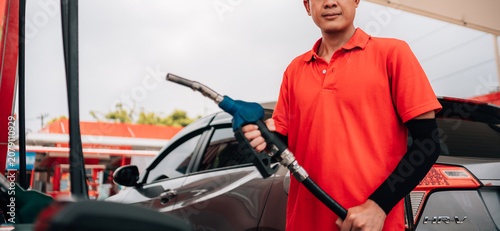 male gas station attendant looking directly at the camera with a confident, approachable expression. He is wearing a polo shirt and a matching cap,he is holding a fuel pump nozzle with a blue handle.