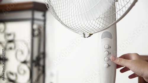 Female hand presses the power button to switch on an electric pedestal fan at home. Close-up of starting a household fan for cooling, ventilation and comfort during hot summer weather.
