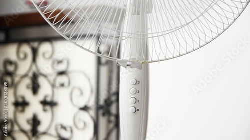Electric pedestal fan operating indoors to provide cooling airflow during hot summer weather. Close-up of a running household fan creating ventilation and improving comfort in a modern home.
