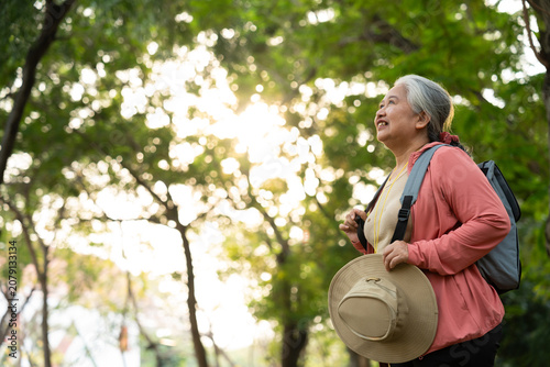 Portrait of Asian mature woman in a hat with backpack behind her back, an Asia active senior woman enjoying nature in autumn park. Standing on a trail in a forest outdoors. Enjoying active travel trip