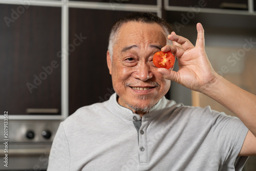 Senior man holding tomato near face in playful concept, symbolizing healthy lifestyle, nutrition, and wellness in cozy indoor kitchen environment with natural daylight, vegetables, and cheerful