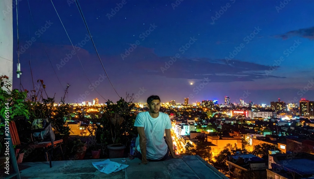 Fototapeta premium Young man in white t-shirt sitting on a rooftop edge gazing at the vibrant night city skyline under a twilight sky with twinkling stars