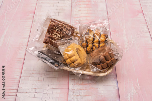 Assorted individually wrapped baked goods, including brownies, cookies, and pastries, arranged in a small woven basket on a rustic pink wooden surface.