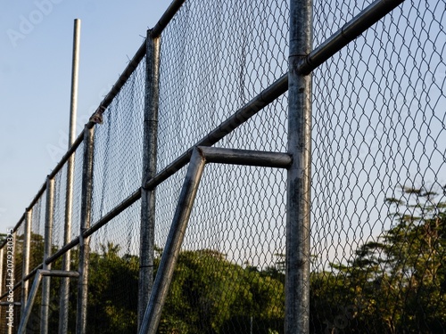 A striking geometric perspective of silver metal grid wire fencing over a lush green tree canopy line.