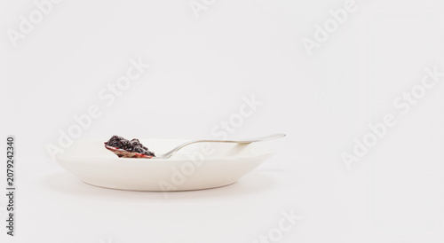 Immagine primo piano con cucchiaio da dessert colmo di confettura di lamponi su un piccolo piatto bianco, sfondo bianco