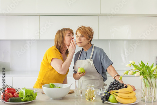 Mid adult  women gossip telling secrets and chatting while cooking in domestic kitchen at home