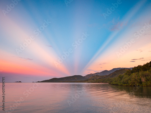 Crepuscular rays fan across the twilight sky over Riung and 17 Islands National Park, Indonesia, illuminating calm coastal waters and lush hills in a serene tropical sunset landscape