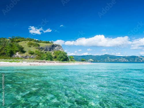 Crystal clear turquoise waters surround a small tropical island in Riung 17 Islands National Park, Indonesia. A peaceful beach, lush green hills, and blue skies create a serene paradise