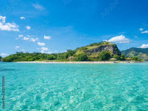 Crystal clear turquoise waters surround a small tropical island in Riung 17 Islands National Park, Indonesia. A peaceful beach, lush green hills, and blue skies create a serene paradise