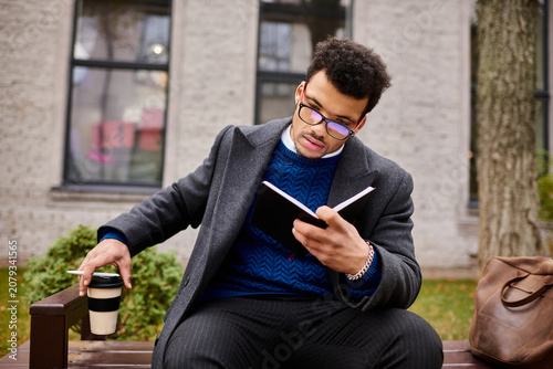 Obraz na plátně African american gentleman absorbed in reading a book, sipping coffee on a sunny day in a serene park