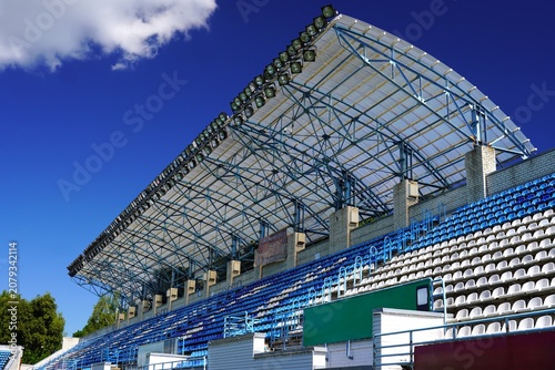  Image of empty stadium grandstands featuring a steel roof structure and blue sky. Rows of blue and white seats are visible, reflecting an organized and geometric design