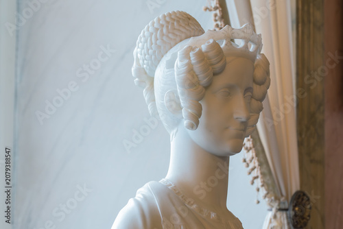 A detailed marble sculpture of a woman's bust, adorned with an intricate headdress and curls, is captured in profile against a softly lit, curtained background, exuding an air of classical elegance an