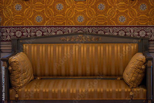 A richly decorated sofa with golden striped upholstery is showcased against a backdrop of intricate, patterned wallpaper, evoking a sense of ornate historical interior design