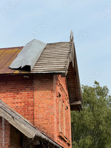 Close-up of a rustic old brick building with wooden roof shingles and a metal sheet under a clear sky, surrounded by greenery