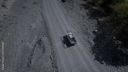 Pickup truck traversing dusty mountain roads with dramatic elevation changes, captured from aerial perspective showing the vehicle navigating through remote natural landscape with rolling hills