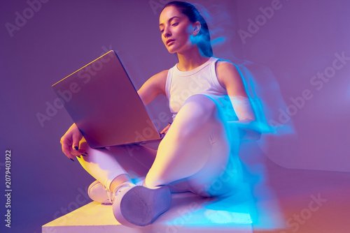 Creative modern young fitness girl works online on laptop, sitting in colorful neon light. Freelance work. Long exposure