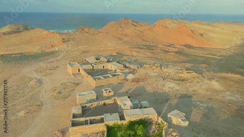 Aerial view of a traditional stone village on Socotra Island, Yemen, surrounded by rugged mountains and the Arabian Sea, illuminated by warm golden hour light in a remote coastal landscape.