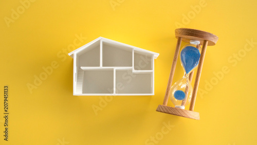 White Plastic Architectural Miniature House Model standing next to Hourglass on Yellow Background
