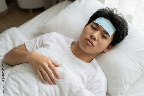 Sick young Asian man lying in bed with cooling gel patch on his forehead, suffering from high fever or flu.