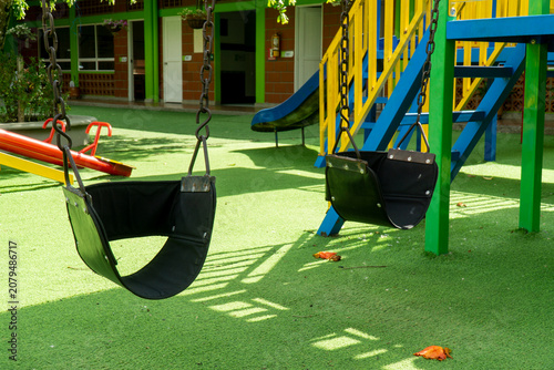 Close up of two empty black rubber swings on chains at an outdoor children playground with colorful structures and artificial grass turf.