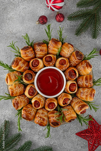 Christmas wreath of pigs in blankets with tomato sauce, garnished with fresh rosemary and sesame. Christmas appetizer on a stone background. Top view, copy space.﻿