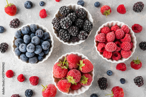 Assorted frozen berries in white bowls: strawberries, blueberries, raspberries, and blackberries on a light stone background. Healthy food. Top view, copy space.