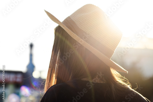 Woman with long hair wearing a straw hat stands outdoors, sunlight illuminating her silhouette against a blurred urban background with buildings and structures