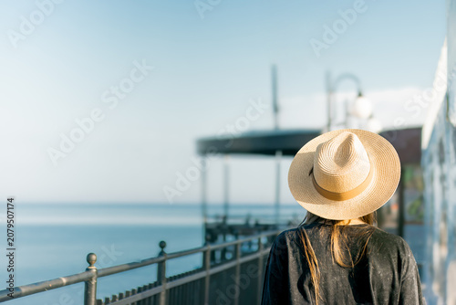 Woman wearing a wide-brimmed straw hat stands by a railing overlooking a calm sea with a clear sky and distant structures visible in the background
