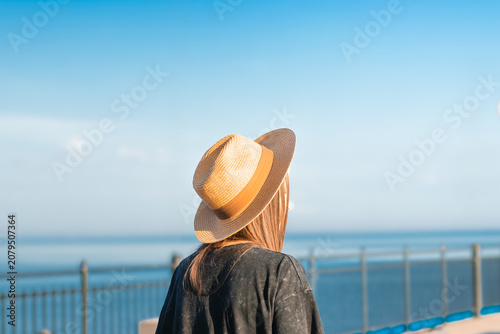 Woman with long brown hair wearing a straw hat stands by a railing overlooking a calm sea under a clear blue sky on a sunny day
