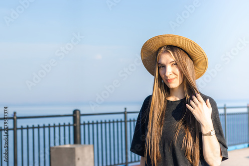 Young woman with long brown hair wearing a straw hat poses by the seaside, with a clear blue sky and calm water in the background