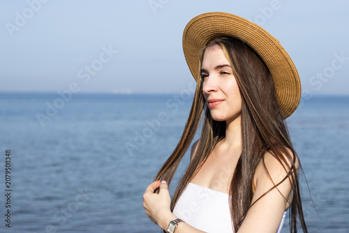 Young woman with long brown hair wearing a straw hat poses by the seaside, with calm blue water and clear sky in the background