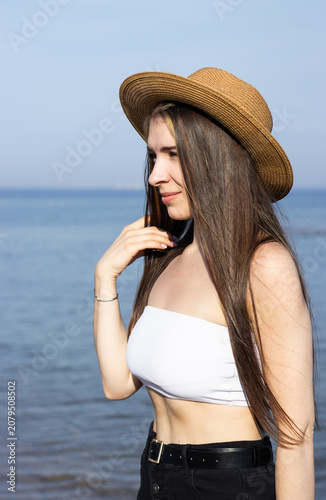 Young woman with long brown hair wearing a straw hat and white crop top poses by the water's edge on a sunny day with a clear blue sky in the background