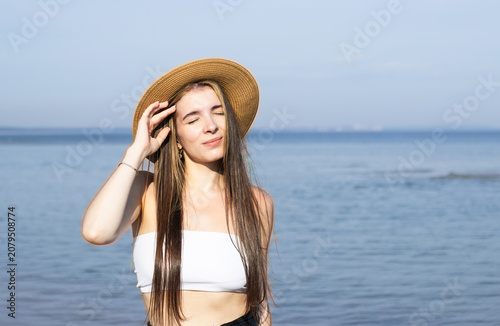 Young woman with long hair wearing a straw hat and white crop top stands by the beach, with calm ocean waves and clear blue sky in the background
