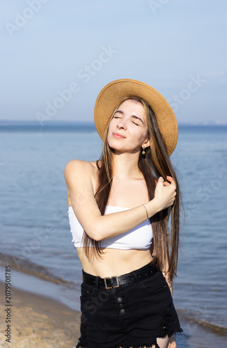 Young woman with long hair wearing a straw hat and white crop top poses by the beach, with calm water and clear sky in the background