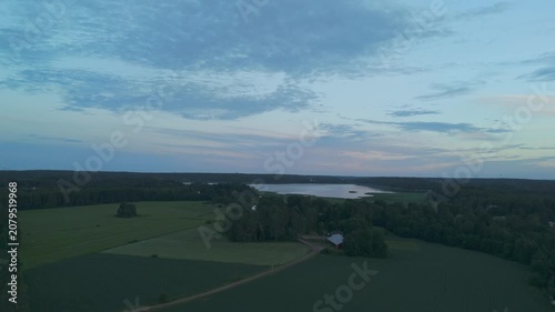 aerial drone flight over green fields toward a forest and peaceful lake with a solitary countryside cabin surrounded by untouched natural landscape