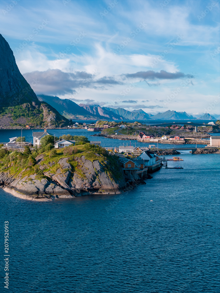 Fototapeta premium Tiny rocky islet in the Lofoten Islands, Norway, with turf roof cabins and rorbuer huts by a small pier, deep fjord and jagged mountains, summer daylight, scattered clouds.