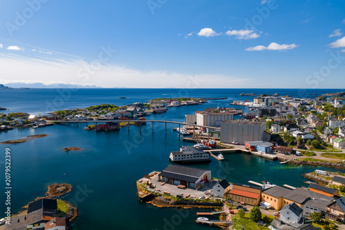 Bild auf Leinwand Aerial view of a Lofoten Islands town in Norway shows a low bridge, islets, rorbuer houses, fisheries, silos, and boats in a sheltered harbor under a clear summer sky
