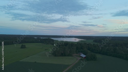 drone flying above open countryside with green fields leading toward a forest lake and a lone cabin surrounded by peaceful wilderness and natural scenery